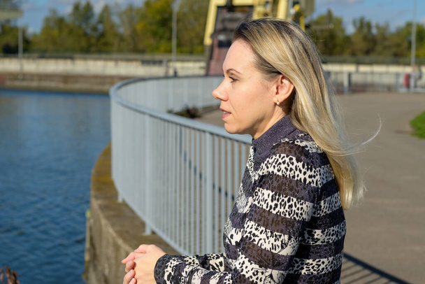 Vista laterale di una donna di mezza età con lunghi capelli biondi si appoggia contro una ringhiera di fronte a una serratura sul Reno in autunno - Foto, immagini