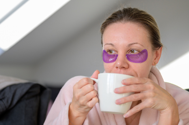 Portrét zamyšlené ženy středního věku v růžovém županu s vycpávkami na oči, popíjející její ranní kávu - Fotografie, Obrázek