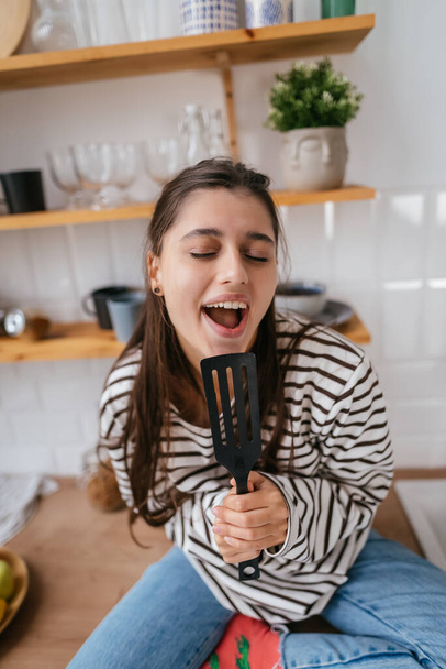Drôle belle femme chantant dans la spatule, tenant la spatule comme microphone, fille ludique s'amusant à la cuisine. - Photo, image