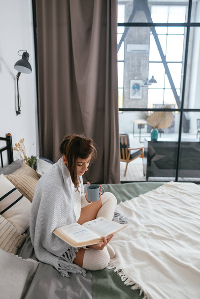Mooie vrouw die boek leest en geniet van warme koffie in zonnig appartement.Ontspanning in bed. - Foto, afbeelding