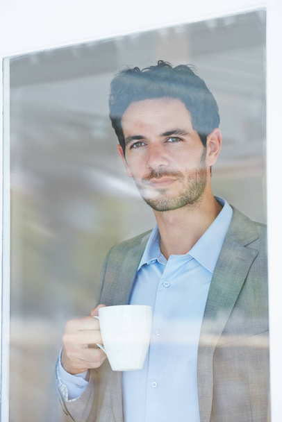 Taking time to enjoy his morning. Portrait of a handsome young man drinking coffee while staring out of a window - Photo, Image
