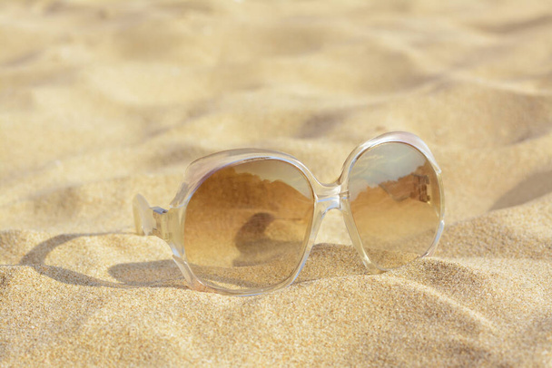Stylish sunglasses with light brown lenses on sandy beach, closeup - Фото, изображение