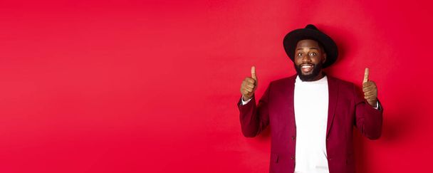 Fashion and party concept. Cheerful african american man in classy jacket, celebrating New Year xmas, showing thumbs up and smiling, like and approve, red background. - Photo, Image