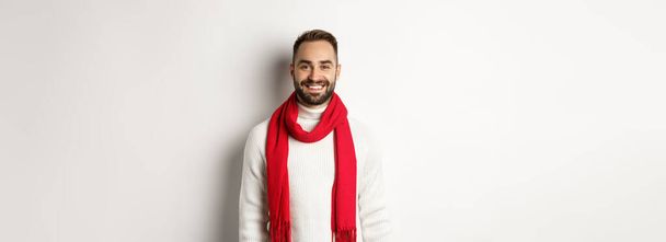 Зимние каникулы. Красивый взрослый мужчина с красным шарфом смотрит счастливо в камеру, стоя в свитере на белом фоне. - Фото, изображение