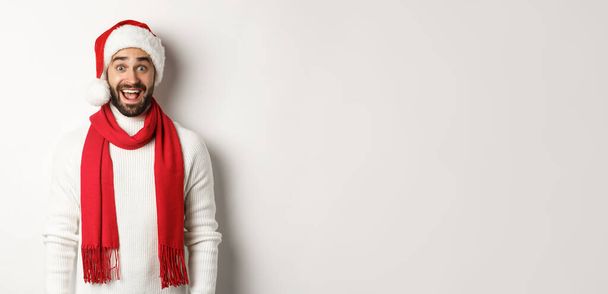 Christmas holidays. Bearded man looking surprised at camera, wearing santa party hat and red scarf, standing against white background. - Foto, imagen