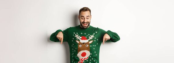 Zimní prázdniny a Vánoce. Šťastný vousatý muž v zeleném svetru, dívá se a ukazuje prsty dolů na reklamu, ukazuje kopírovací prostor, bílé pozadí. - Fotografie, Obrázek