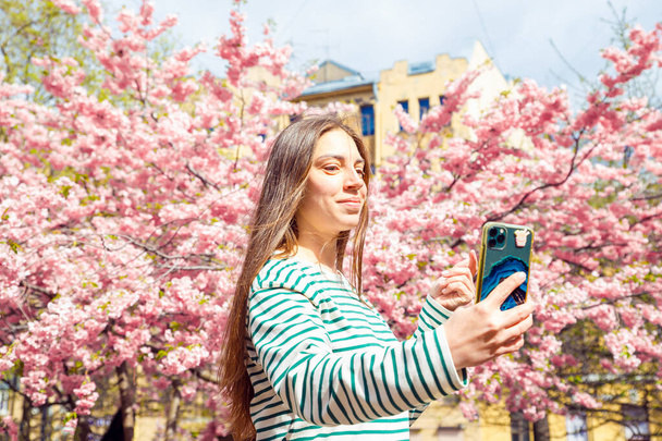Portrait faisant selfie femelle sur smartphone sur cerisier fleur arbre sakura. Jeune femme caucasienne souriante prend selfie portrait avec téléphone mobile. Été Printemps tour de taille mode de vie portrait. - Photo, image