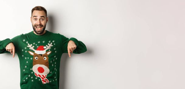 Téli ünnepek és karácsony. Izgalmas szakállas férfi zöld pulóverben, ujjal lefelé mutogatva, csodálkozva, fehér háttér fölött állva. Fénymásolási hely - Fotó, kép