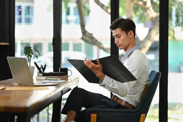 Handsome zakenman met behulp van laptop en het doen van wat papierwerk op helder modern kantoor. - Foto, afbeelding