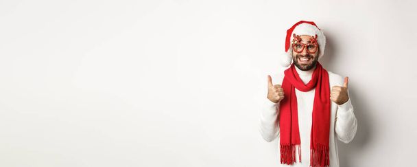 Navidad, Año Nuevo y concepto de celebración. Hombre excitado en Santa sombrero y gafas de fiesta, mostrando los pulgares hacia arriba en la aprobación, sonriendo satisfecho, fondo blanco. - Foto, Imagen
