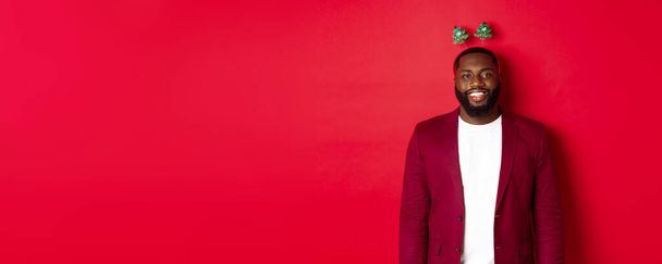 Joyeux Noël. Heureux homme afro-américain célébrant le Nouvel An, portant bandeau de fête drôle et souriant, debout sur fond rouge. - Photo, image