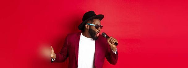 Gepassioneerde zwarte mannelijke zanger optredend tegen rode achtergrond, zingen in microfoon, het dragen van party outfit, staande over rode achtergrond. - Foto, afbeelding