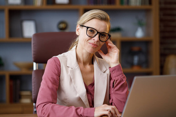 Portrait of beautiful blondie woman looking at camera and smiling. Adult stylish confident businesswoman wearing eyeglasses sitting at workplace in office. Portrait of modern successful female leader. - Foto, imagen
