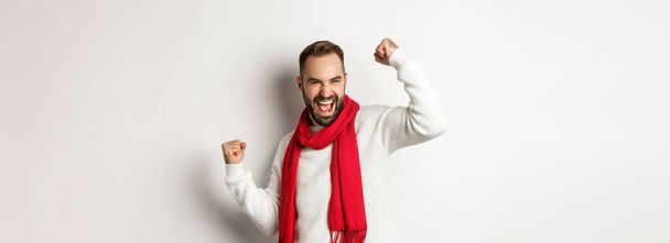 Christmas holidays and New Year concept. Happy bearded man celebrating and enjoying party, raising hands up in triumph, rejoicing while standing over white background. - Fotó, kép