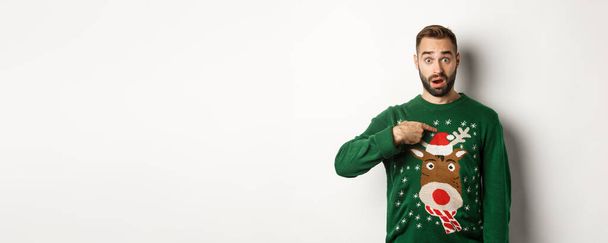 Téli ünnepek és karácsony. Összezavarodott szakállas fickó, aki magára mutat, megijed az ajánlattól, fehér háttér fölött áll pulóverben.. - Fotó, kép