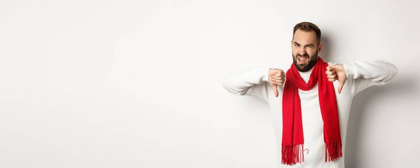 Sceptische en ontevreden man met duimen naar beneden, het dragen van winter trui met sjaal, een hekel aan een oneens, staande over witte achtergrond. - Foto, afbeelding