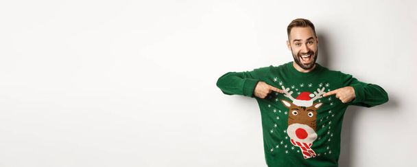 Nieuwjaarsviering en wintervakantie concept. Vrolijke man wijzend naar zijn grappige kerst trui en glimlachende, witte achtergrond. - Foto, afbeelding