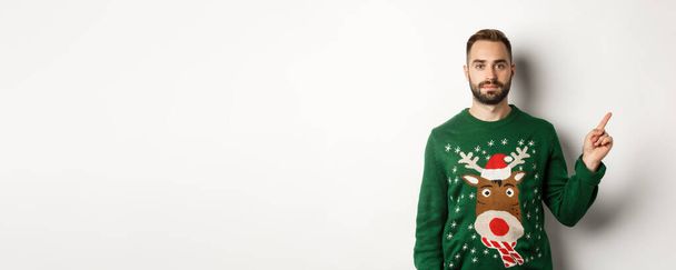 Vacaciones de invierno y Navidad. Hombre barbudo guapo en suéter verde apuntando con el dedo hacia la derecha, mostrando promo, de pie sobre fondo blanco. - Foto, imagen