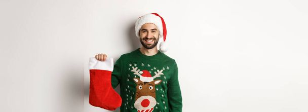 Концепция рождественской вечеринки и праздников. Счастливый человек в шляпе Санты приносит подарки в рождественском носке и улыбается, стоя на белом фоне. - Фото, изображение