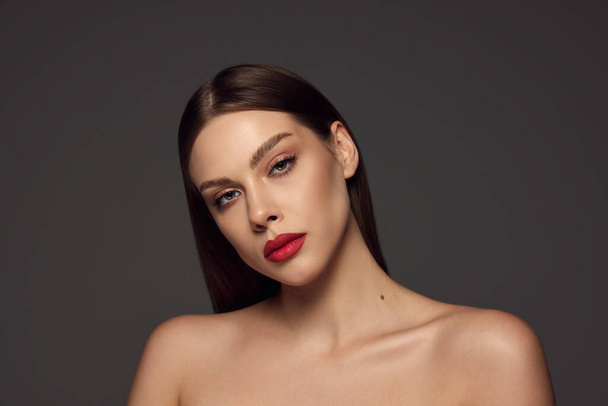 Female fashion model with naked shoulders and long hair looking at camera over dark grey background. Beautiful woman with trendy make-up, red lipstick and well-kept skin. Magazine style, beauty. - 写真・画像
