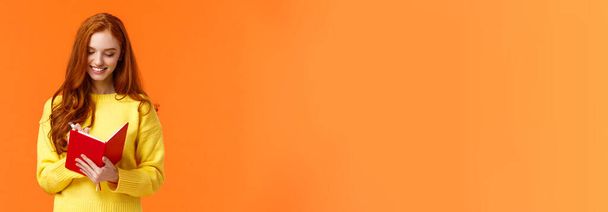 Taille-up portret aantrekkelijke roodharige meisje twintiger jaren schrijven in haar dagboek, in te vullen to-do lijst of persoonlijke schema in nieuwe notebook, glimlachen alsof alles gepland, staande oranje achtergrond zorgeloos. - Foto, afbeelding