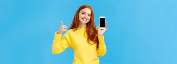 Girl showing good app, recommend download very useful application. Cute redhead woman in yellow sweater, like winter holiday sale in online store, showing smartphone display, blue background. - Фото, изображение