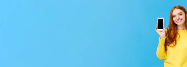 Chica mostrando buena aplicación, recomiendo descargar aplicación muy útil. Mujer pelirroja linda en suéter amarillo, como la venta de vacaciones de invierno en la tienda en línea, mostrando la pantalla del teléfono inteligente, fondo azul
. - Foto, imagen