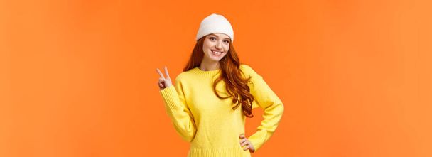 Nowoczesny hipster dziewczyna gra śnieżki z przyjaciółmi podczas ferii zimowych, pokazując znak pokoju jako pozowanie zdjęcie w pobliżu bałwana, noszenie białej czapki i sweter na pomarańczowym tle. - Zdjęcie, obraz