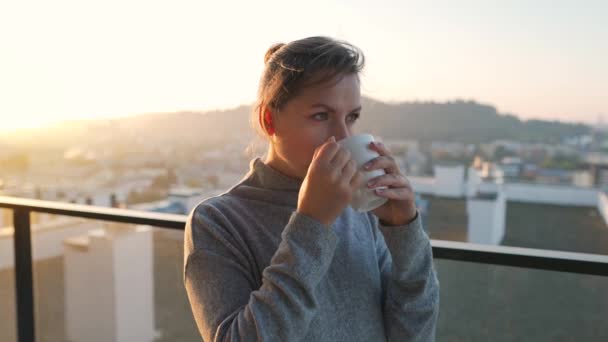 Vrouw begint haar dag met een kopje thee of koffie op het balkon bij dageraad, slow motion. Stadslandschap op de achtergrond. Moderne stedelijke levensstijl - Video