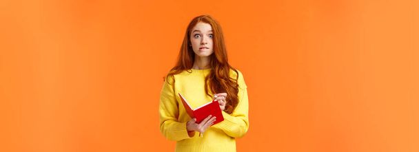 Problemen roodharige blanke meisje vergeten afspraak, herinneren taak na het kijken notebook, bijten lip en staren bezorgd of beschaamd, het opschrijven van belangrijke notities, oranje muur. - Foto, afbeelding