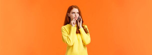 besluiteloze en onzekere, sceptische ernstig uitziende roodharige vrouw die moeilijke beslissingen neemt tijdens telefoongesprek, fronsende aanraking lip pensive, bestelling levering van voedsel, oranje achtergrond. - Foto, afbeelding
