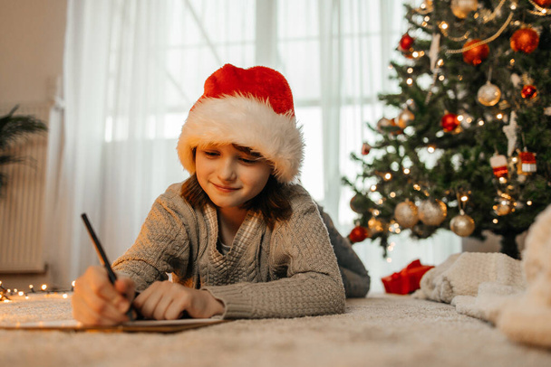 Újév és Karácsony. Egy tinédzser lány télapó sapkában levelet ír a Mikulásnak egy díszített karácsonyfa mellé koszorúban és fényben.. - Fotó, kép