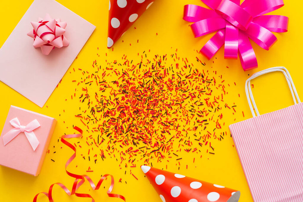 Top view of festive gift near party caps and sprinkles on yellow background  - Foto, Imagen