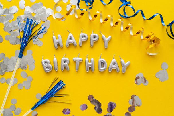 Top view of candles in shape of Happy Birthday lettering near confetti and serpentine on yellow background  - Foto, immagini