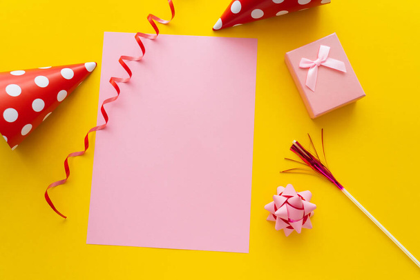 Vue du dessus de la carte de vœux vide près des casquettes de fête en pointillés et de la boîte cadeau sur fond jaune  - Photo, image