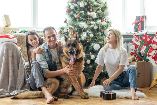 Família amigável está brincando com o cão perto da árvore de Natal. Eles estão sentados e rindo
. - Foto, Imagem