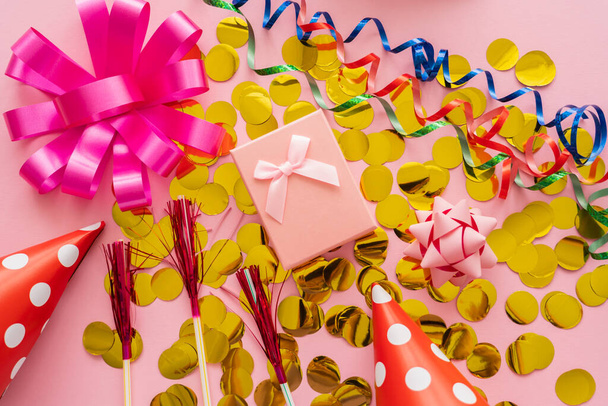 Vue du dessus du présent sur confettis près des chapeaux de fête et serpentine sur fond rose  - Photo, image