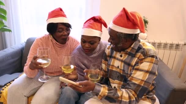 Familia afroamericana de tres personas tomando una selfie con smartphone celebrando el brindis navideño con vino blanco y sombreros de Santa - Metraje, vídeo