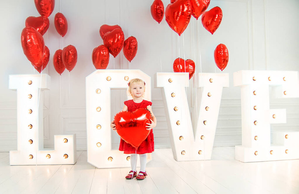 Koncepcja Walentynek. dziewczynka w czerwonej sukience w pobliżu dużych liter napis miłości i z czerwonymi kulkami w postaci serc. Słowo miłość. - Zdjęcie, obraz