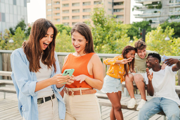 Two attractive and cheerful smiling women looking and showing the last funny and entertaining news on their smartphone outside at the university campus. At background, three young friends, having fun - Photo, Image
