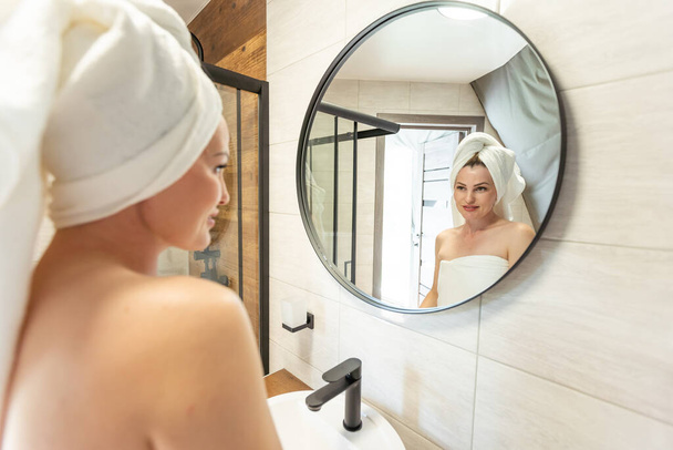 Después del cuerpo de la ducha y la cabeza envuelta en toalla 35s mujer se ve en el espejo toca la piel hidratada suave y saludable de la cara se siente satisfecha disfruta del procedimiento de tratamiento de cosméticos de spa, concepto de higiene del cuidado matutino
 - Foto, imagen