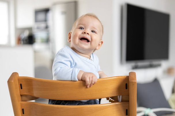 Szczęśliwe niemowlę siedzi w tradycyjnym skandynawskim projektancie drewnianym wysokim krześle i śmieje się głośno w nowoczesnym, jasnym domu. Słodki uśmiech dziecka - Zdjęcie, obraz