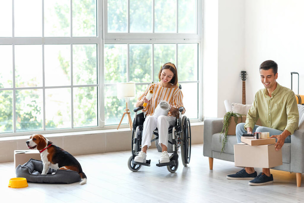 Νεαρός με τη γυναίκα του σε αναπηρική καρέκλα και το σκυλί συσκευασίας πράγματα στο δωμάτιο την ημέρα της μετακίνησης - Φωτογραφία, εικόνα