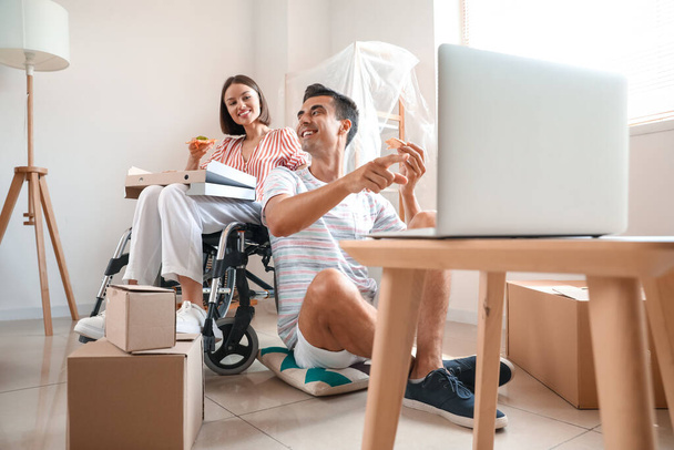 Young man and his wife in wheelchair with pizza watching video in room on moving day - Φωτογραφία, εικόνα