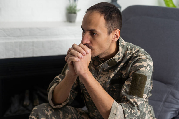 Thoughtful military man staring aside, holding palms by mouth, sitting on couch at home. Young soldier visiting psychologist, suffering from posttraumatic stress, closeup photo, copy space - Foto, Bild