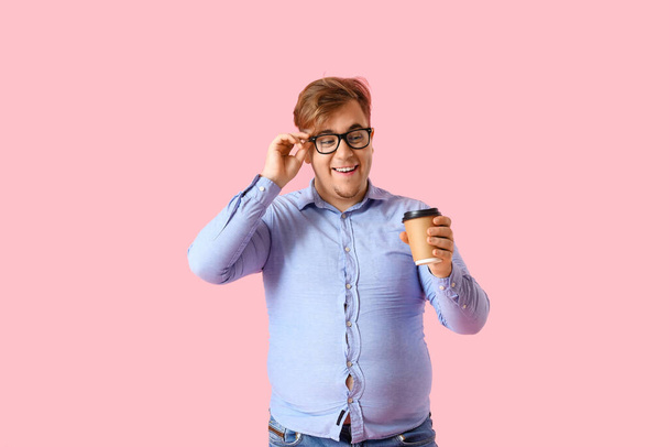 Young overweight man in tight shirt with cup of coffee on pink background - Photo, Image