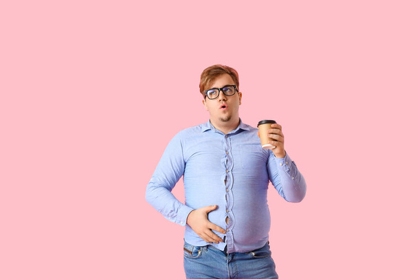 Young overweight man in tight shirt with cup of coffee on pink background - Photo, Image