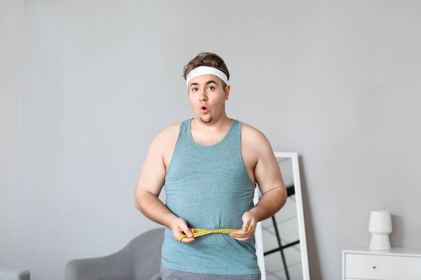 Σοκαρισμένος νεαρός υπέρβαρος άντρας με μεζούρα στο σπίτι - Φωτογραφία, εικόνα