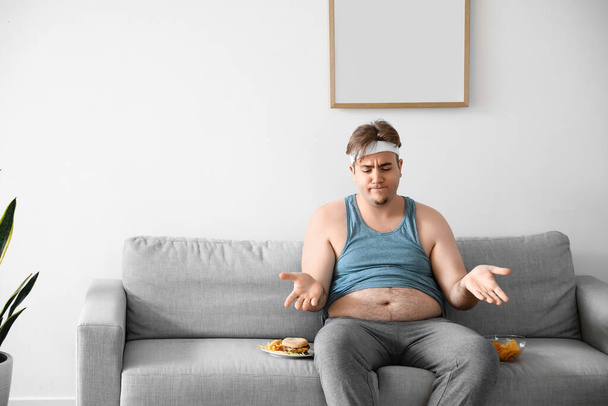 Enojado joven hombre con sobrepeso con comida poco saludable sentado en el sofá en casa - Foto, Imagen