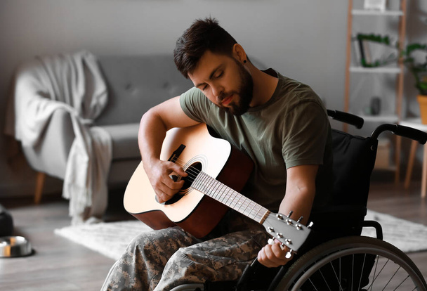 Joven soldado en silla de ruedas tocando la guitarra en casa - Foto, Imagen